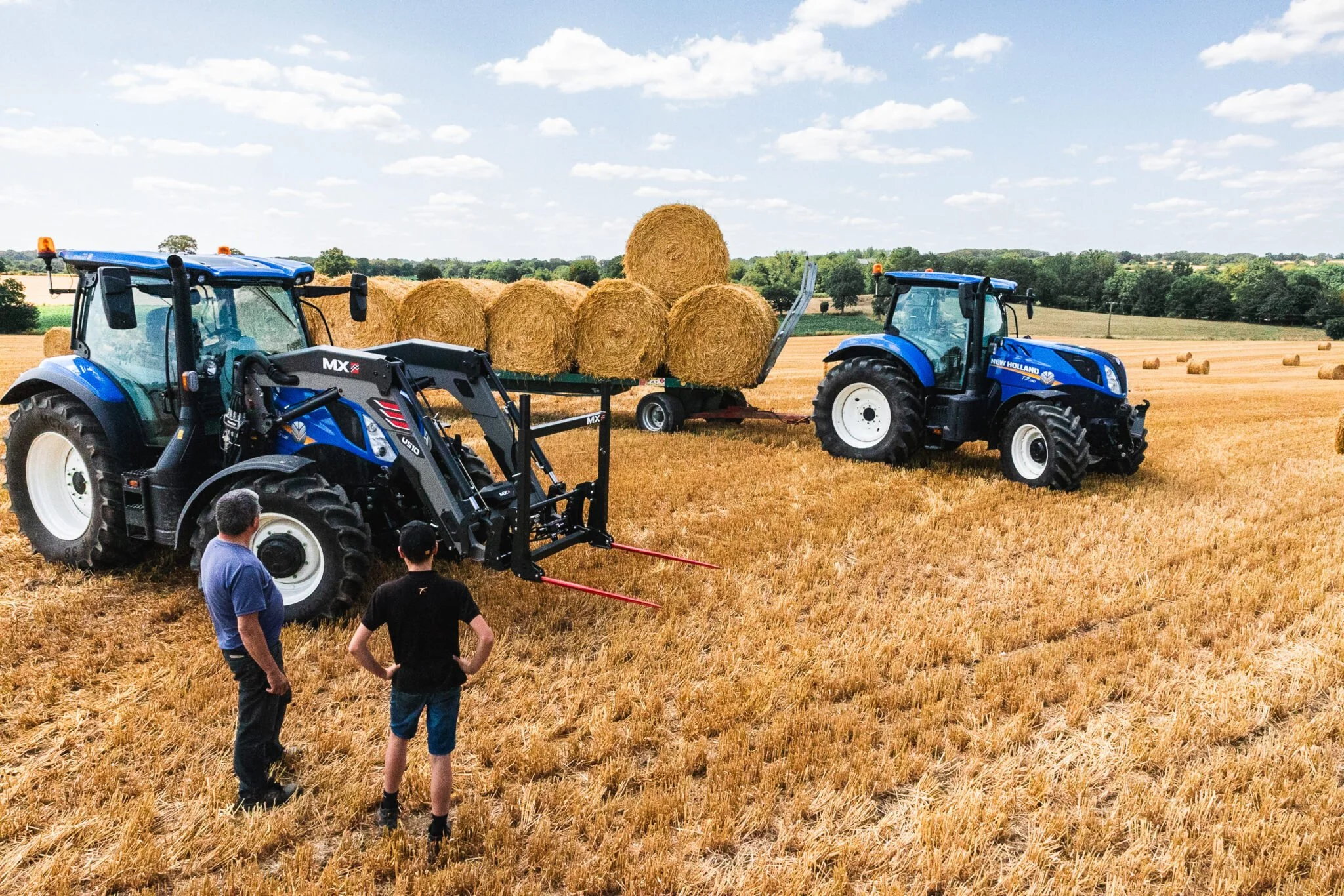 Two blue tractors in a field with hay bales; two people are standing nearby, one man and one boy, observing.