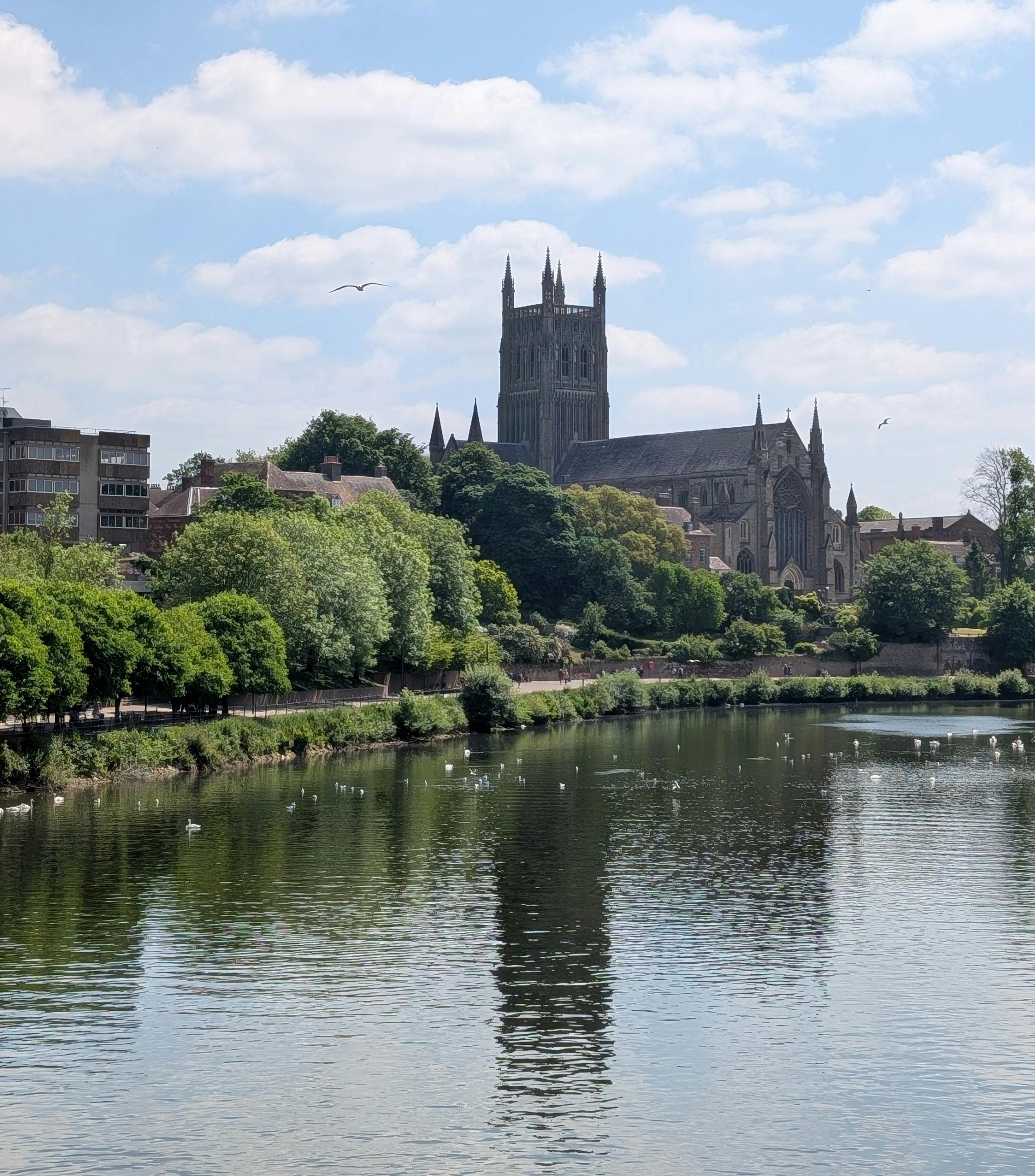 View of a historic cathedral with tall spires overlooking a river, surrounded by green trees and a partly cloudy sky.
