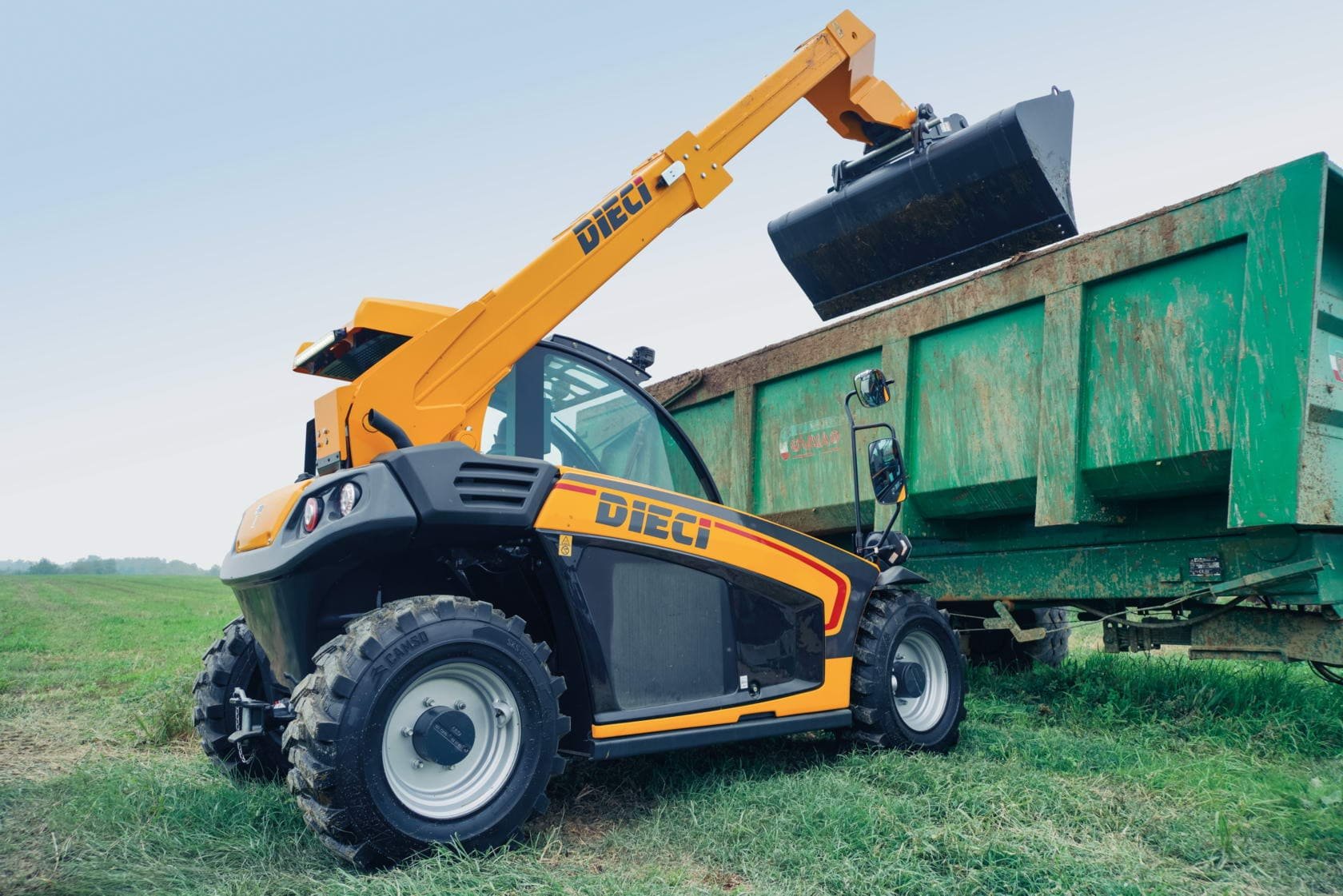 A yellow and black Dieci telehandler lifting a black bucket to dump material into a green trailer on a grassy field.