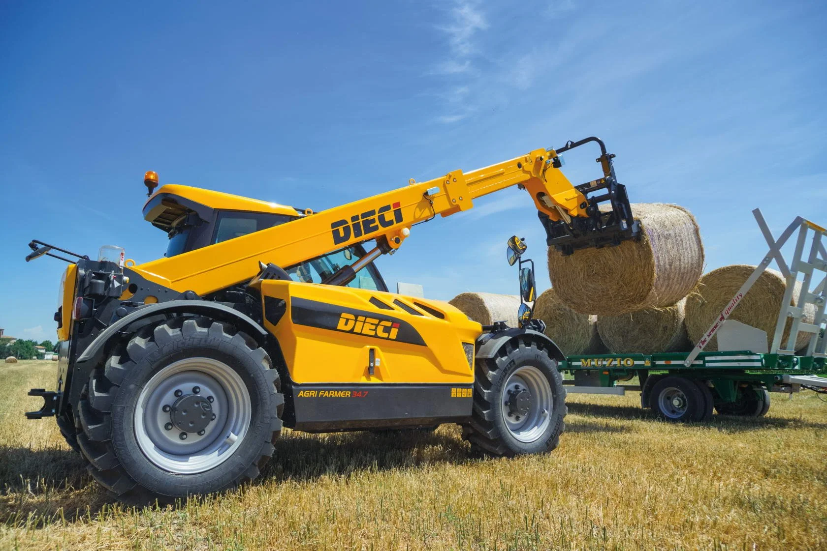 Yellow telescopic loader lifting hay bales onto a green trailer in a field under a blue sky.