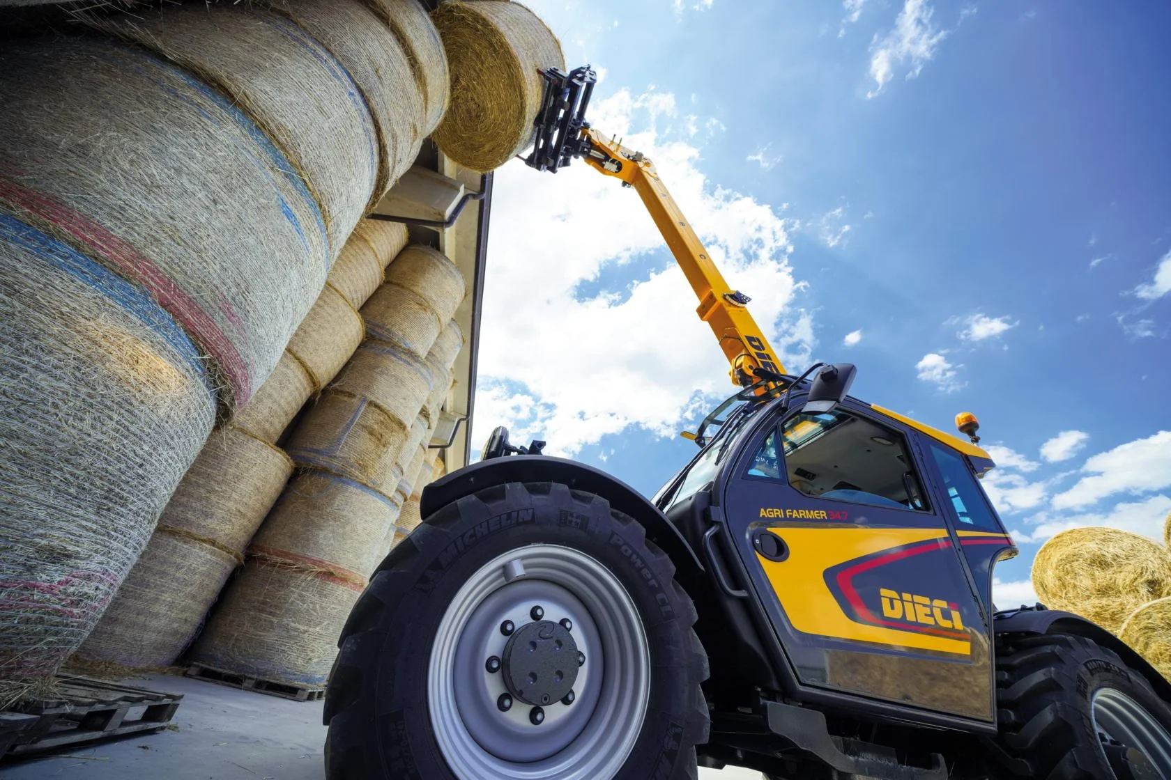 A yellow telehandler with the brand name DIECI lifting hay bales in a farm setting under a partly cloudy sky.