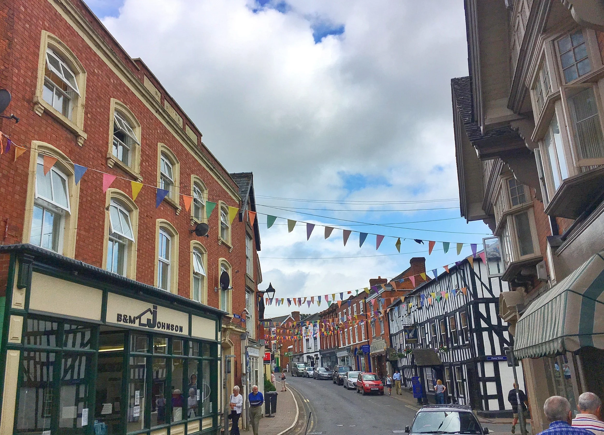 Street view with multi-story buildings, colorful bunting flags across, people walking, parked cars, and a partly cloudy sky.