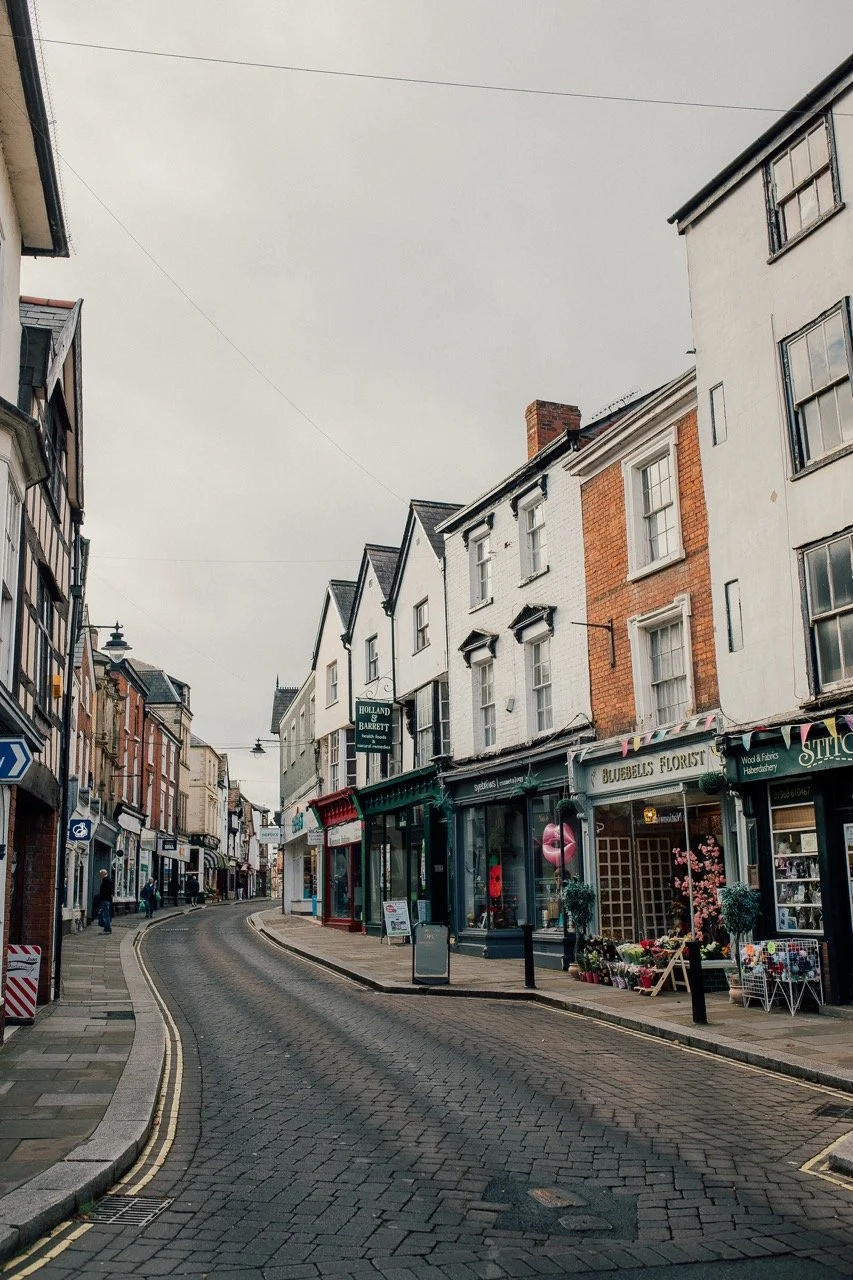 A quiet, curved street lined with shops and buildings in a small town or city, with overcast sky overhead.