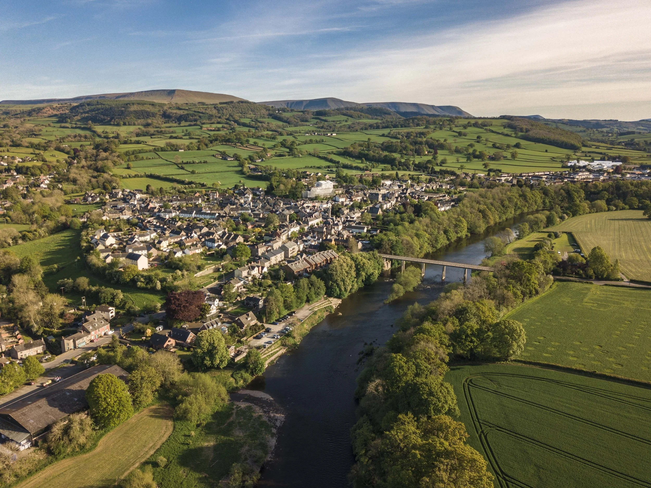 An aerial view of a small town situated along a winding river with green fields and rolling hills in the background, under a mostly clear sky.