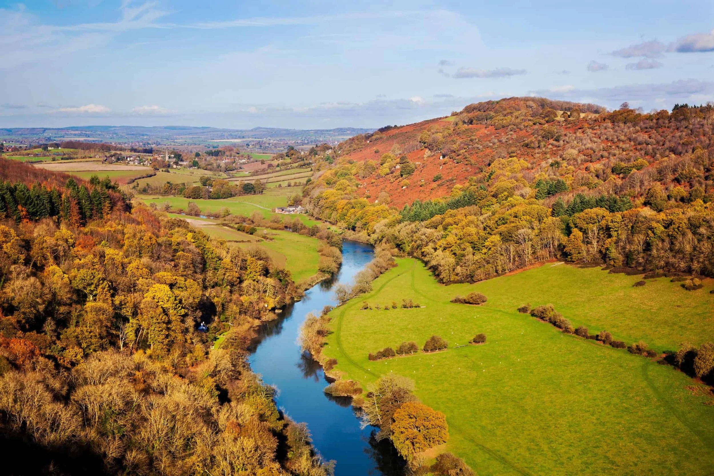 A scenic landscape of a river winding through green fields and forested hills, with a distant view of a small town or village under a partly cloudy sky.