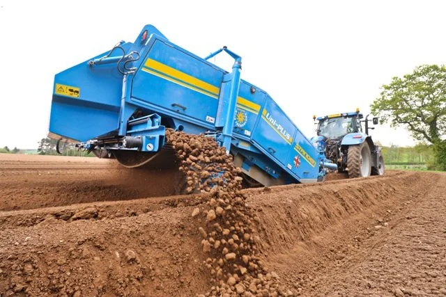 A blue tractor with a large attached soil screen spreading soil and stones onto a farm field.