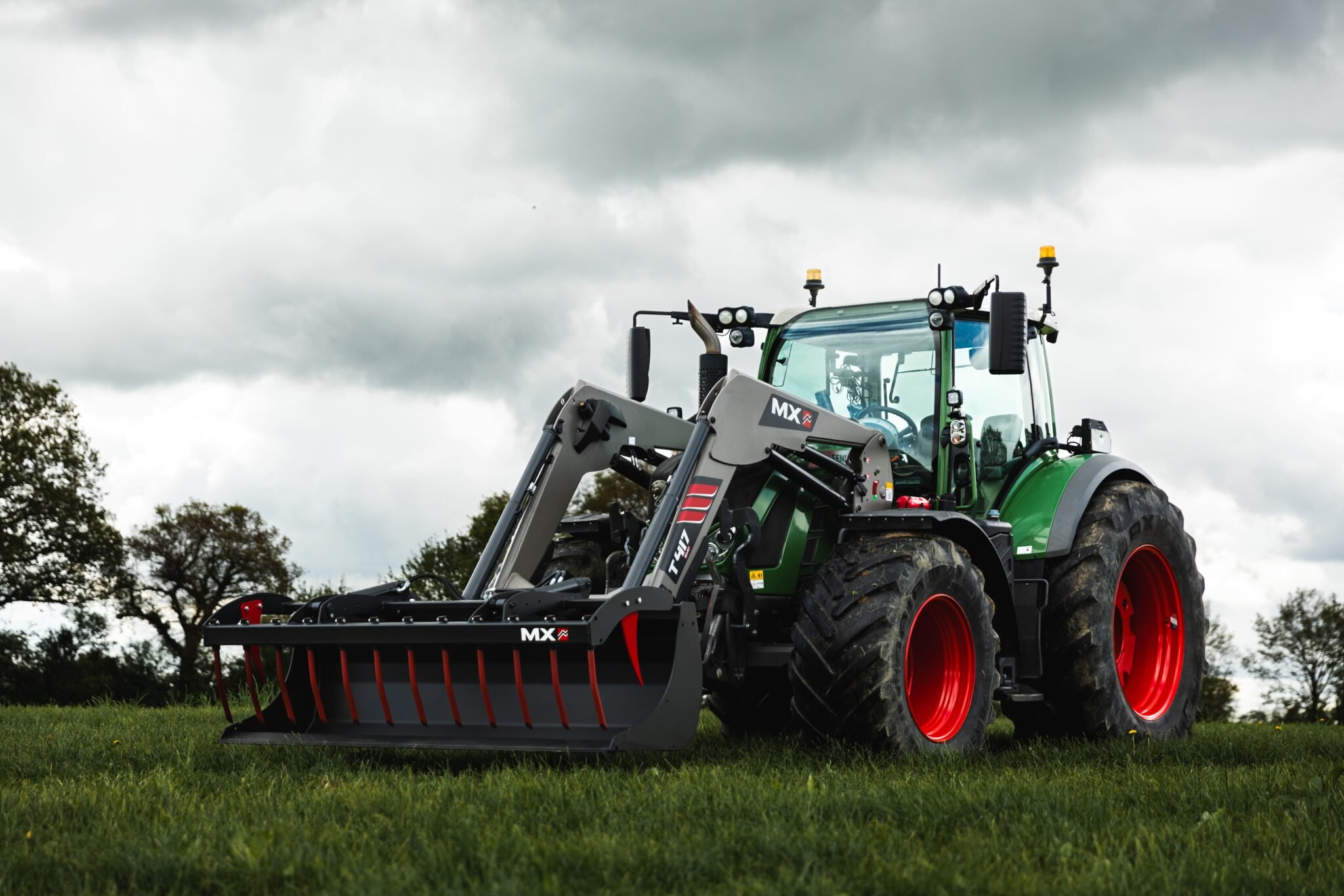 A green tractor with large black tires and red rims on a grassy field, equipped with a front loader attachment against a cloudy sky.