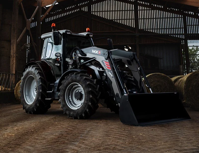 A black tractor with a front loader attachment inside a barn with hay bales.