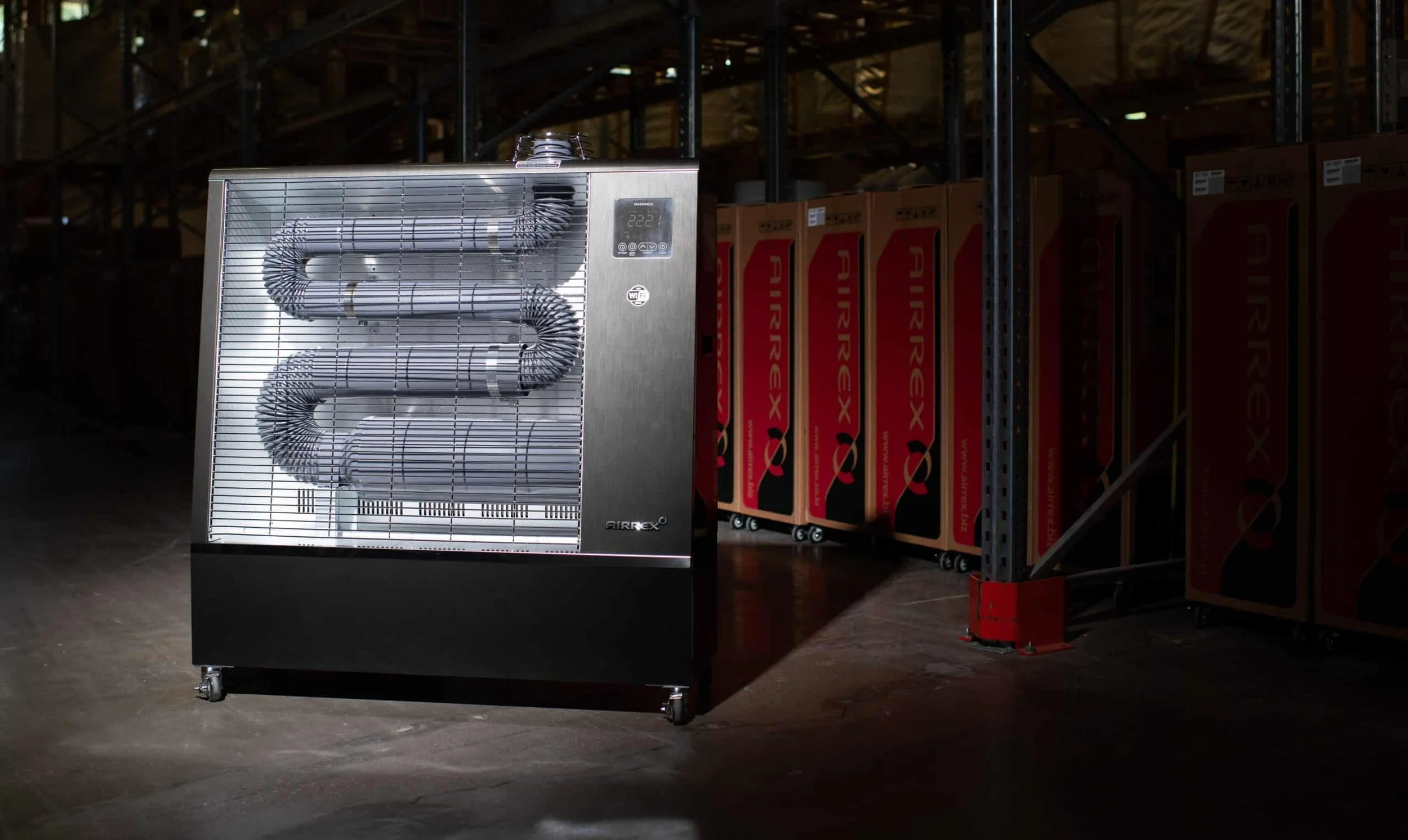 A large commercial space heater with metal grates covering the heating elements, placed on wheels in a warehouse or storage area. In the background, there are stacked boxes and metal shelving racks.