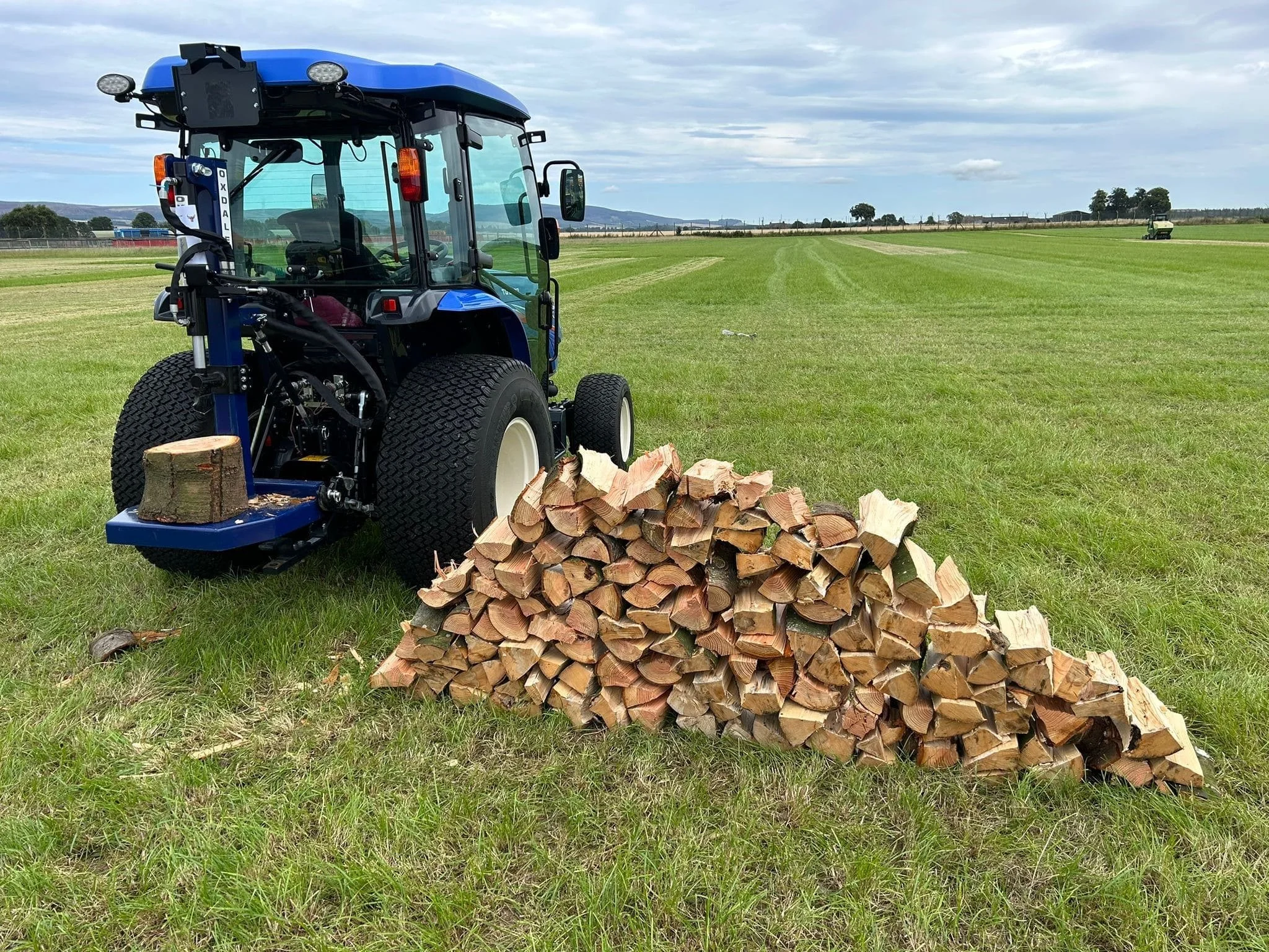 Blue tractor with a pile of chopped firewood on grassy field under cloudy sky.