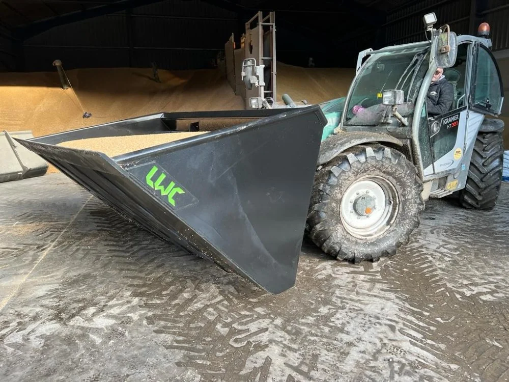 A compact utility loader with a front-mounted snow blower attachment in an indoor storage or warehouse area, with piles of grain or sand in the background.
