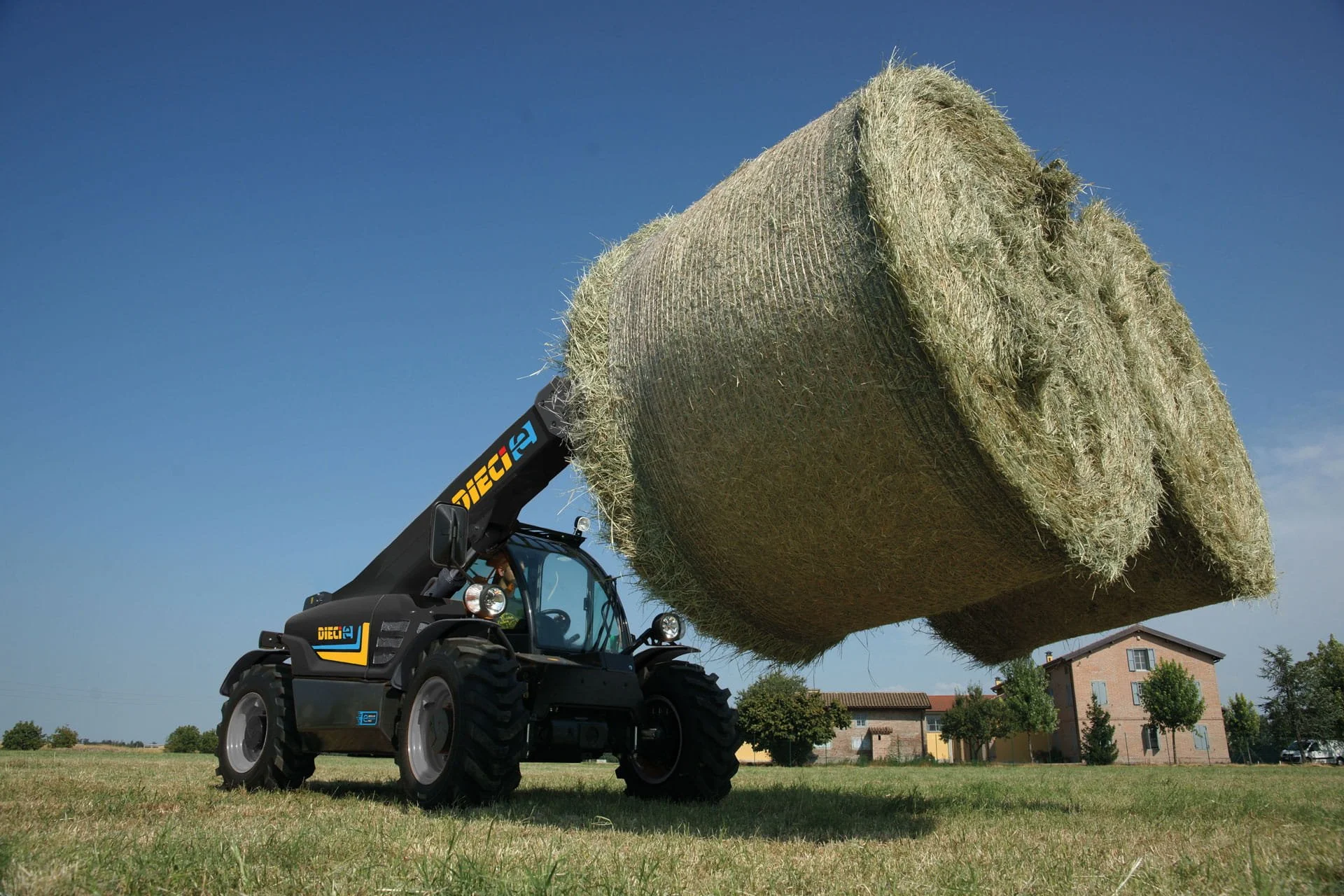 A tractor lifting a large round hay bale in a field under a clear blue sky.