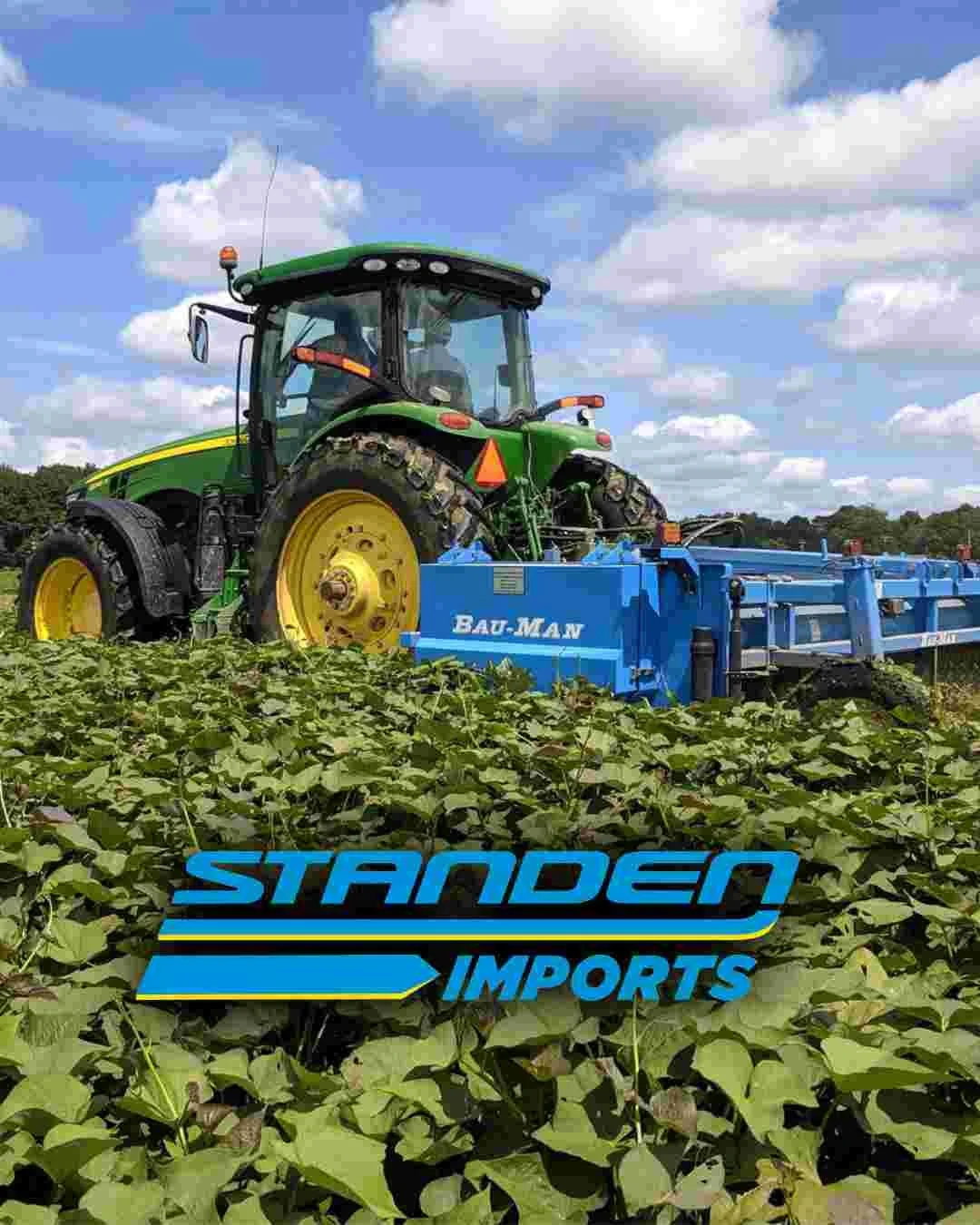 A green and yellow tractor with a blue attachment working in a farm field, with green plants in the foreground and a partly cloudy sky in the background.