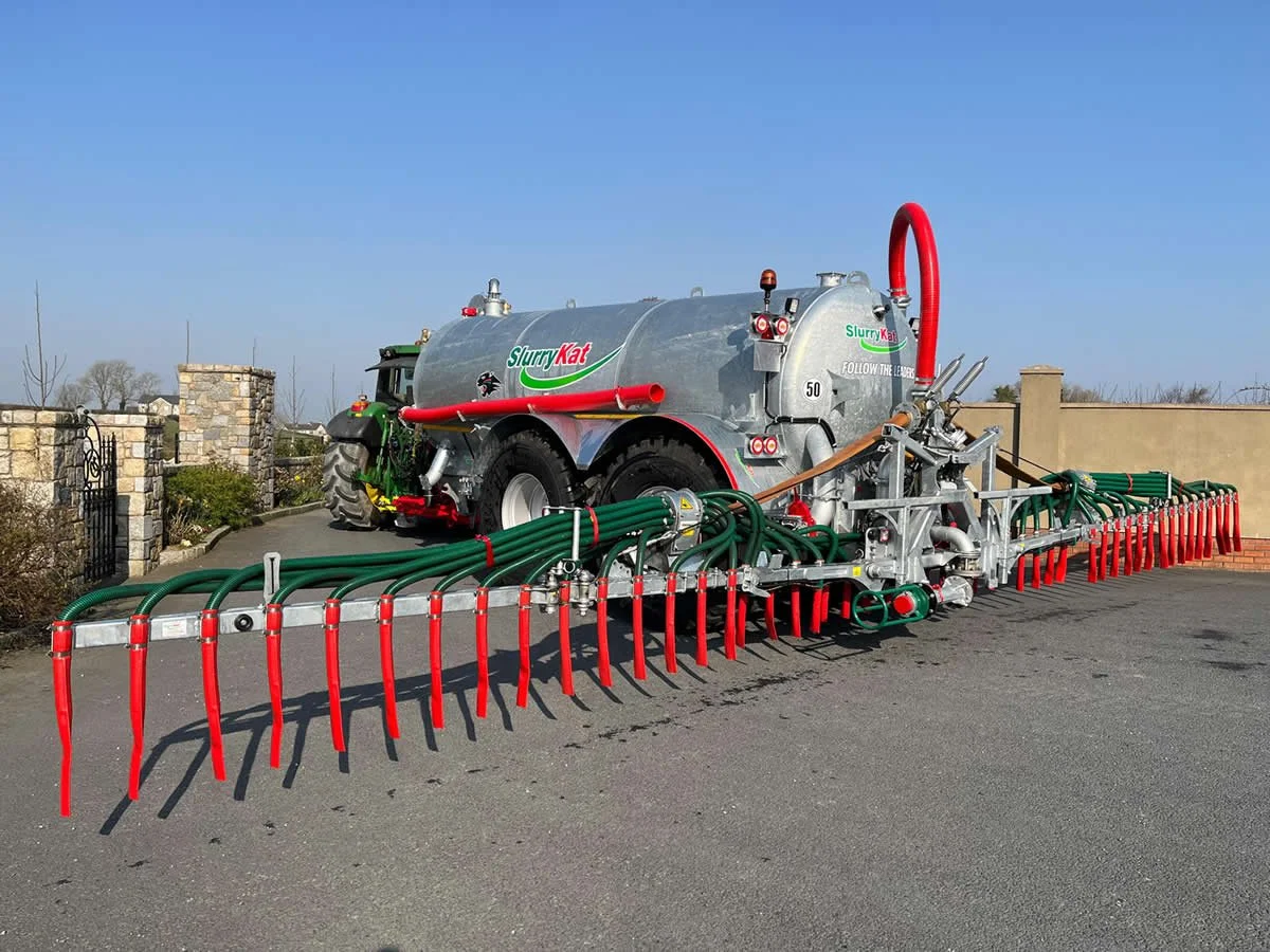 Large roadside agricultural slurry tanker with green hoses and red tines attached for spreading liquid manure or fertilizer on fields, parked on an asphalt surface with a stone wall and fencing in the background.