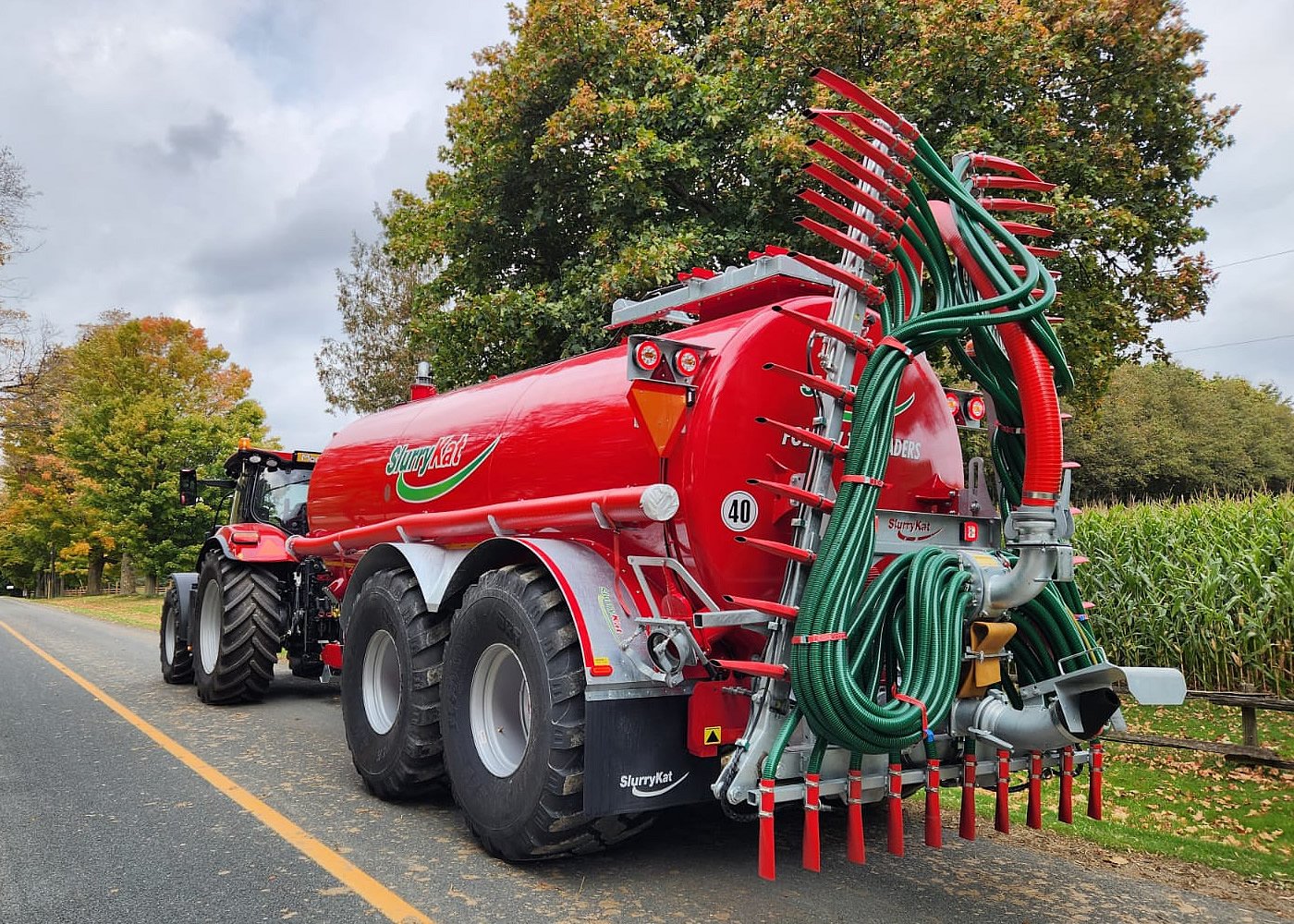 A large red slurry tanker attached to a tractor, with green and red hoses hanging from the back, parked on a rural road with trees and a cornfield in the background.