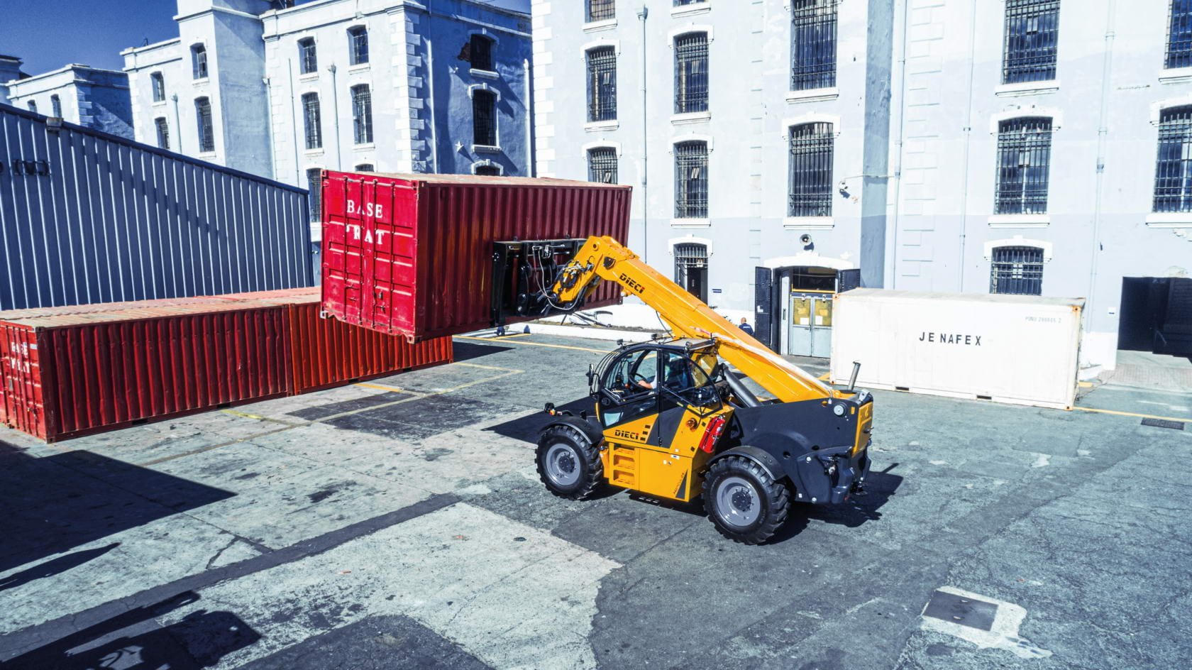 A yellow industrial vehicle lifting red shipping containers in an urban lot with large white buildings in the background.