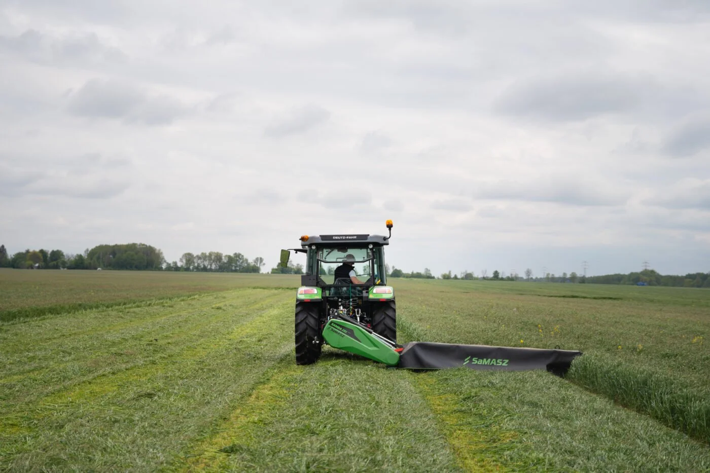 A person using a green tractor with a mower attachment on a grassy field under cloudy skies.