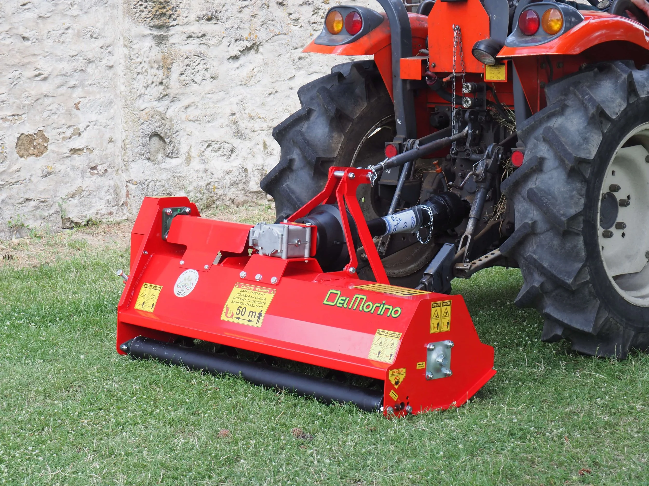 A red tractor attached to a red hay baler in front of a stone wall. The hay baler has caution and warning labels, and the tractor has large black tires.