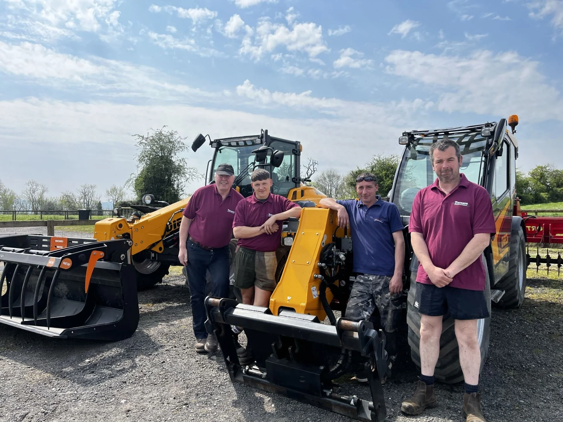 Group of four men standing next to yellow construction equipment outdoors on a gravel surface, with trees and a blue sky with clouds in the background.