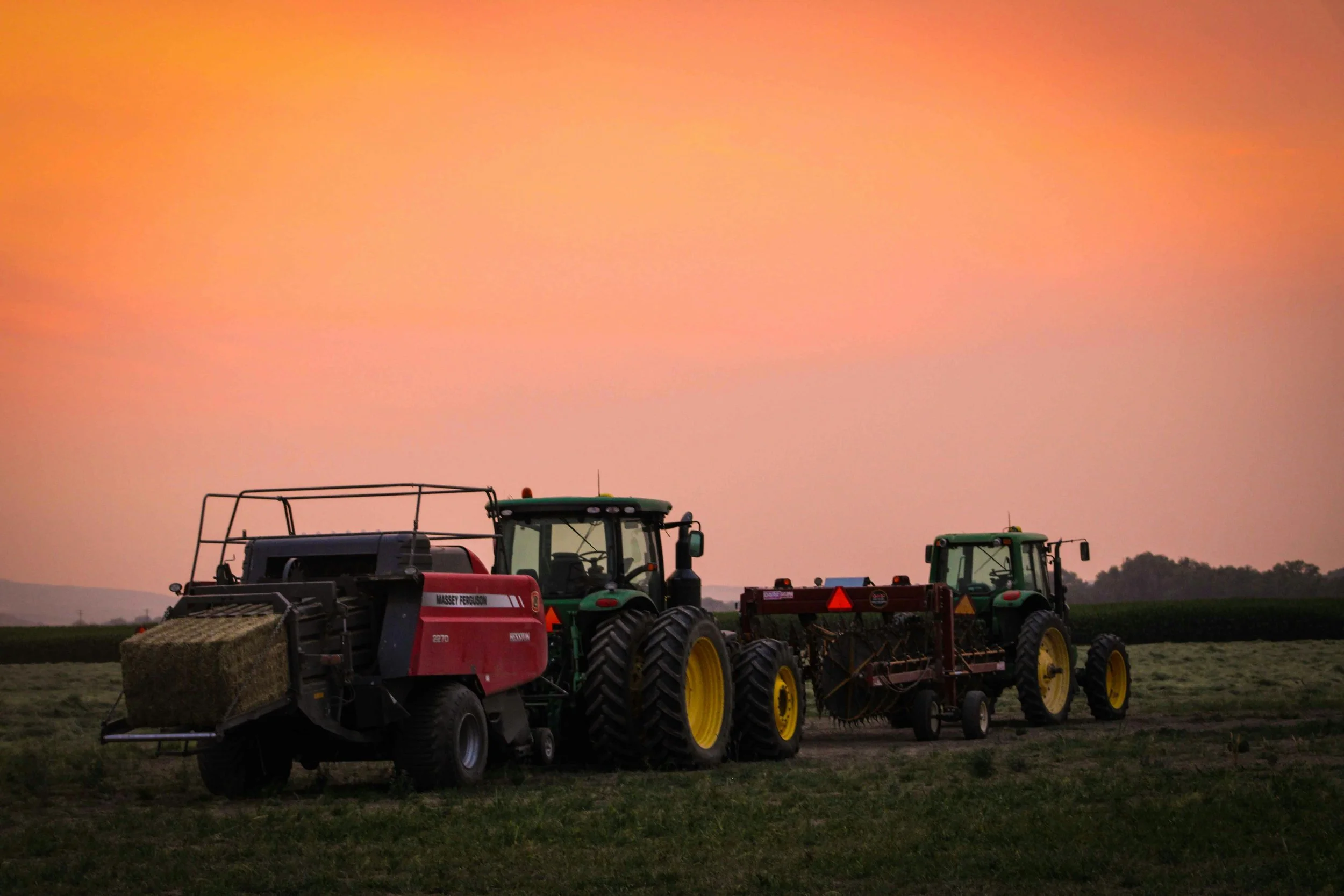 Two tractors working in a field during sunset, one with a bale of hay and the other attached to farming equipment.
