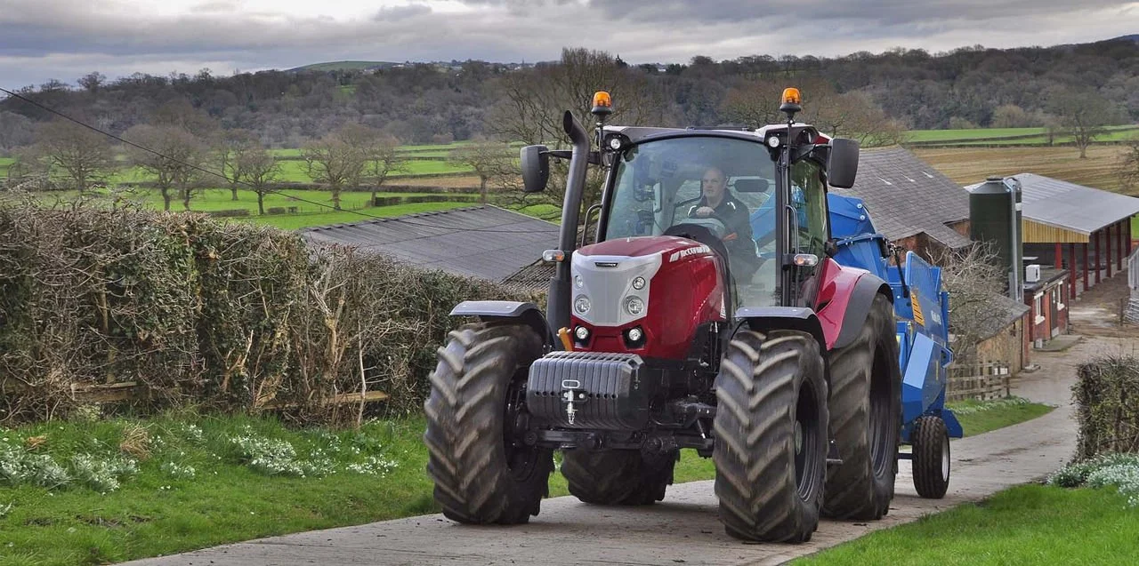 A red tractor pulls a blue agricultural implement along a rural farm road, with fields and barns in the background under an overcast sky.