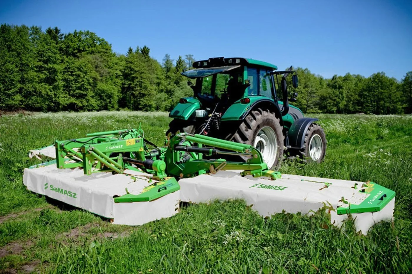Green tractor with attached mowing equipment on a grassy field under a blue sky, surrounded by trees.