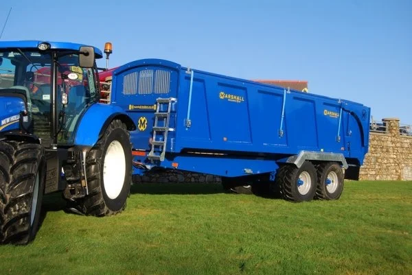Blue tractor with a large attached blue Marshal sweep loader on a grassy field under a clear blue sky.