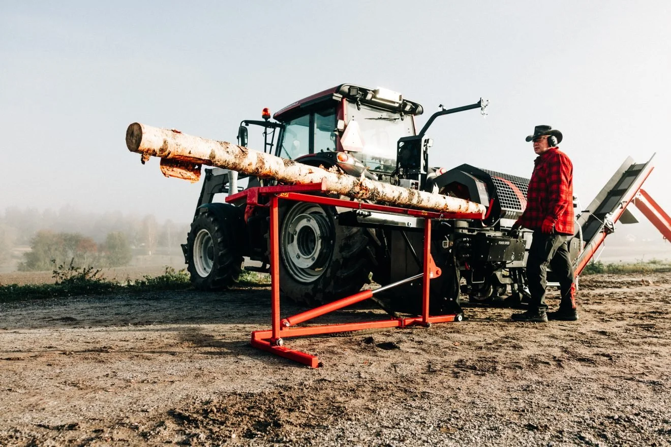 A person standing next to a tree logging machine on a dirt field, with a large log on the machine's conveyor.