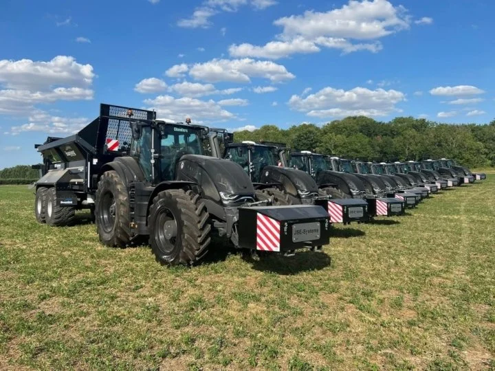 A line of large black tractors parked on grass under a blue sky with clouds.