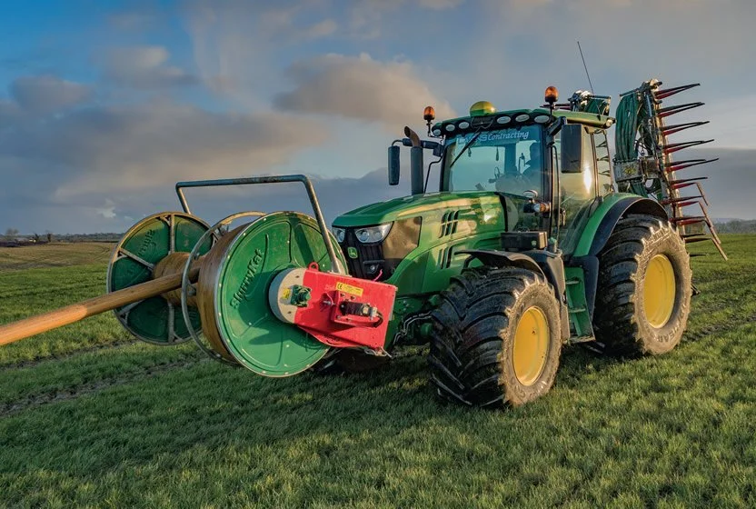 A green John Deere tractor with a large reel and sprayer attachment working on a grassy field during daytime.