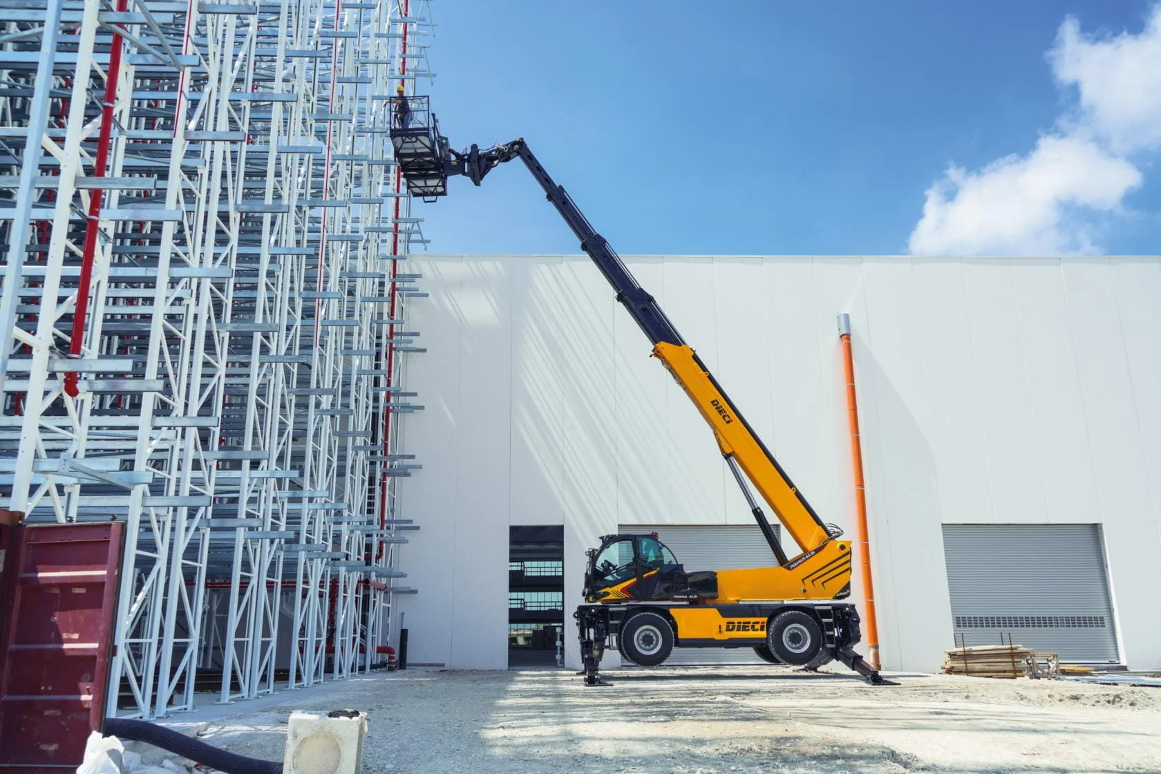 Construction site with scaffolding on the left and a crane lifting a worker on a platform near the scaffolding, against a blue sky with clouds.