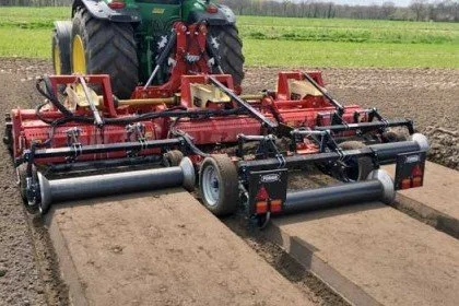 A green tractor towing a red landscape rake attachment on a farm field.