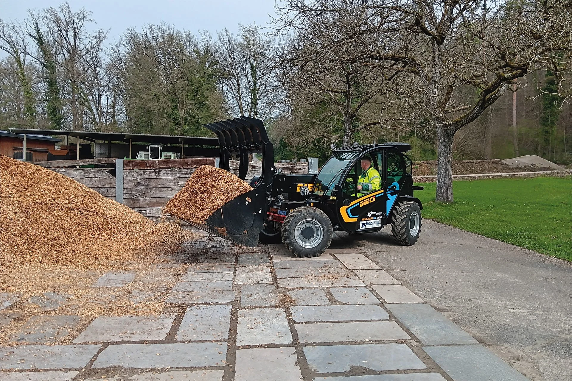 A person operating a small black front-end loader with a yellow and black branding logo, moving wood chips or mulch in an outdoor area with trees and a path.
