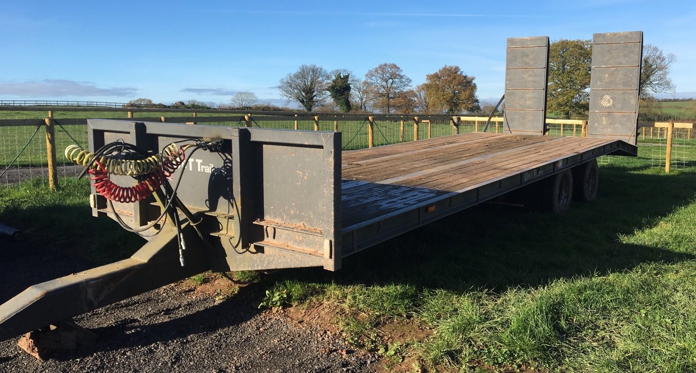 Empty flatbed trailer with wooden floor on grass field, surrounded by a fence, under a clear blue sky.