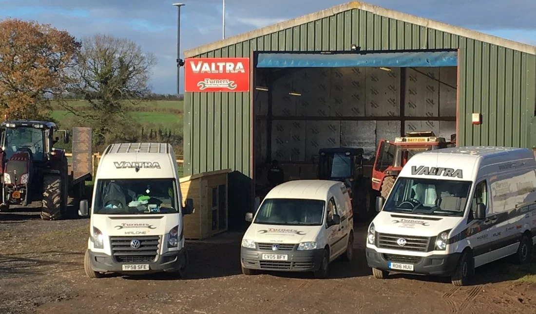 Farm equipment and vehicles parked outside a green metal building with a red Valtra sign, including two Valtra branded vans, a small van, and a tractor on a trailer.