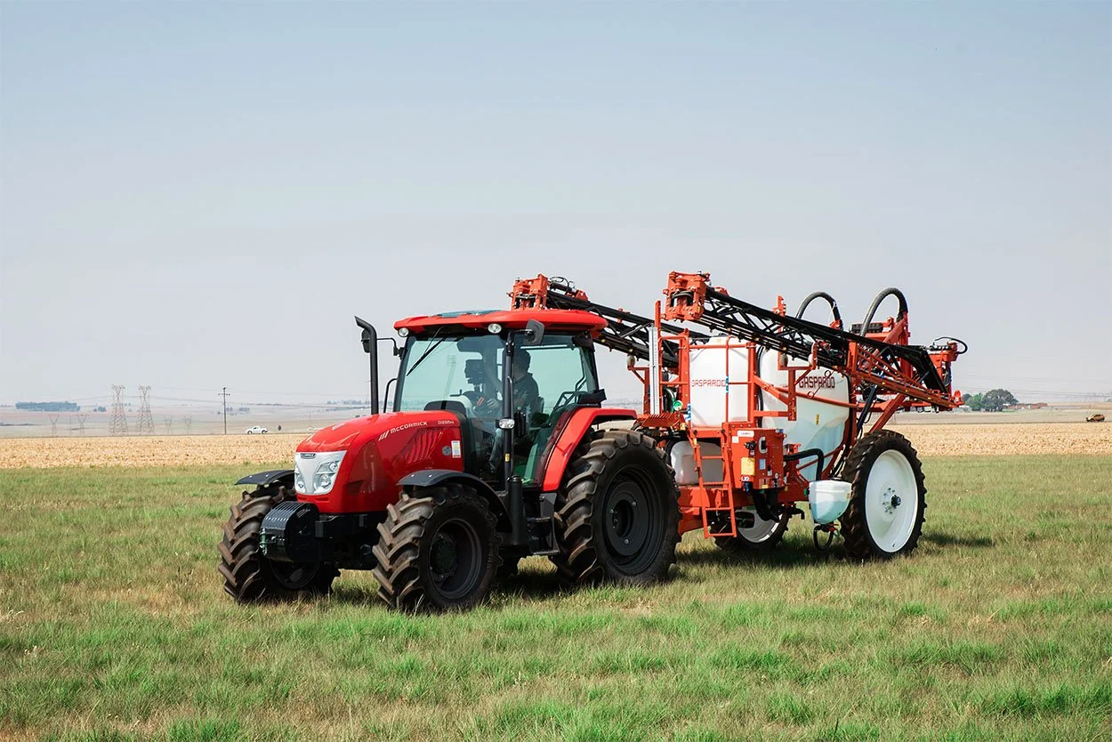A red tractor with a large spray tank attached, working in a green field on a clear day.