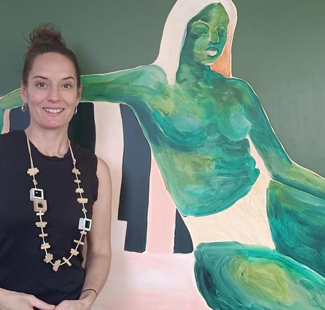 A woman with dark hair tied up, wearing a black sleeveless top and a large necklace, standing next to a painted image of a reclining woman with green and blue tones on the wall.