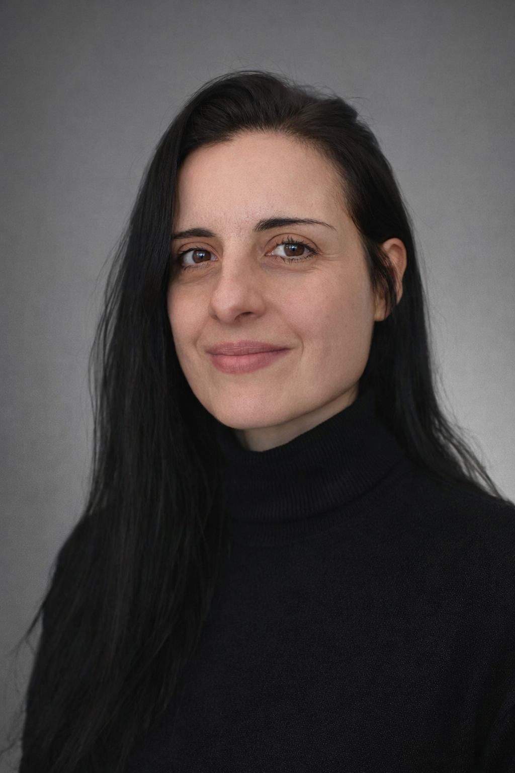 Close-up portrait of a woman with long black hair, wearing a black turtleneck, smiling slightly, against a neutral gray background.