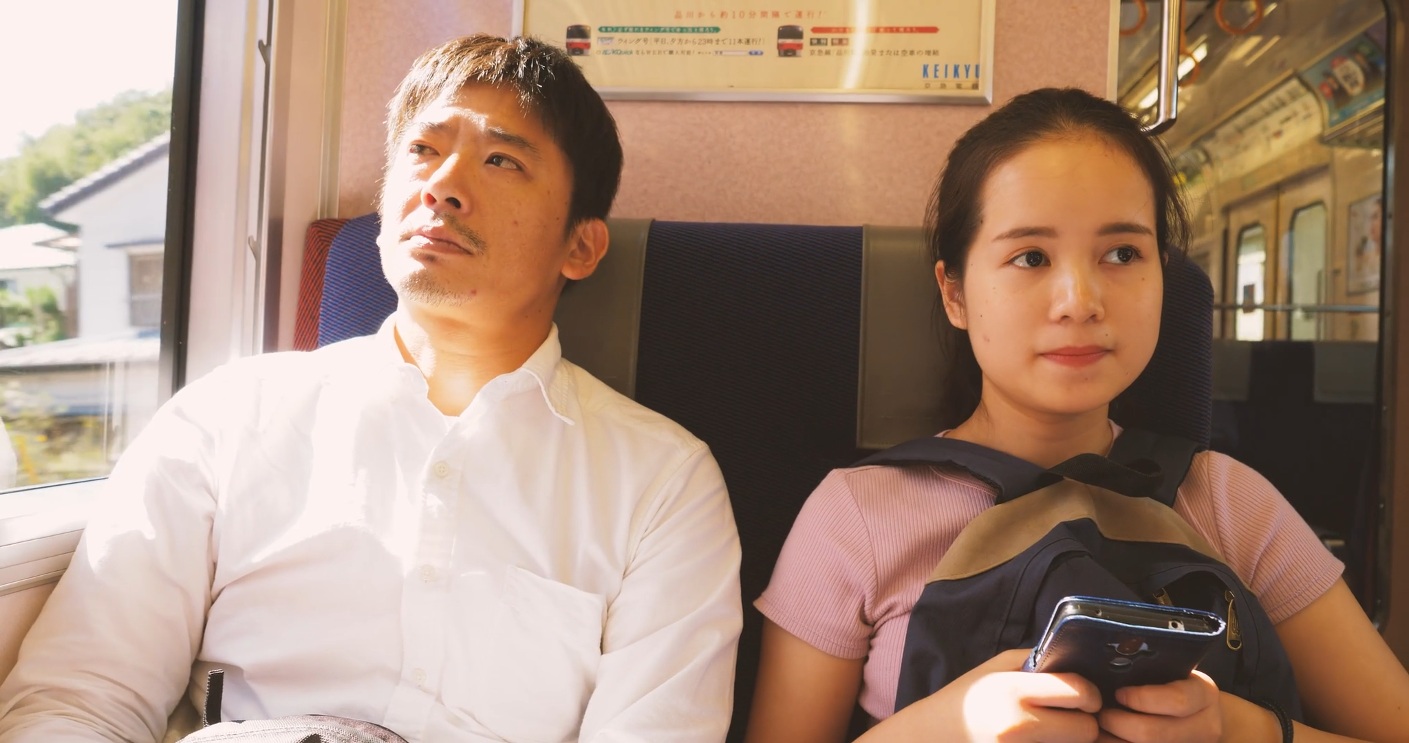 電車の座席に座る男性と若い女性、男性は窓の外を見ていて、女性はスマートフォンを持ちながら座っている。背景に車内と外の景色が見える。