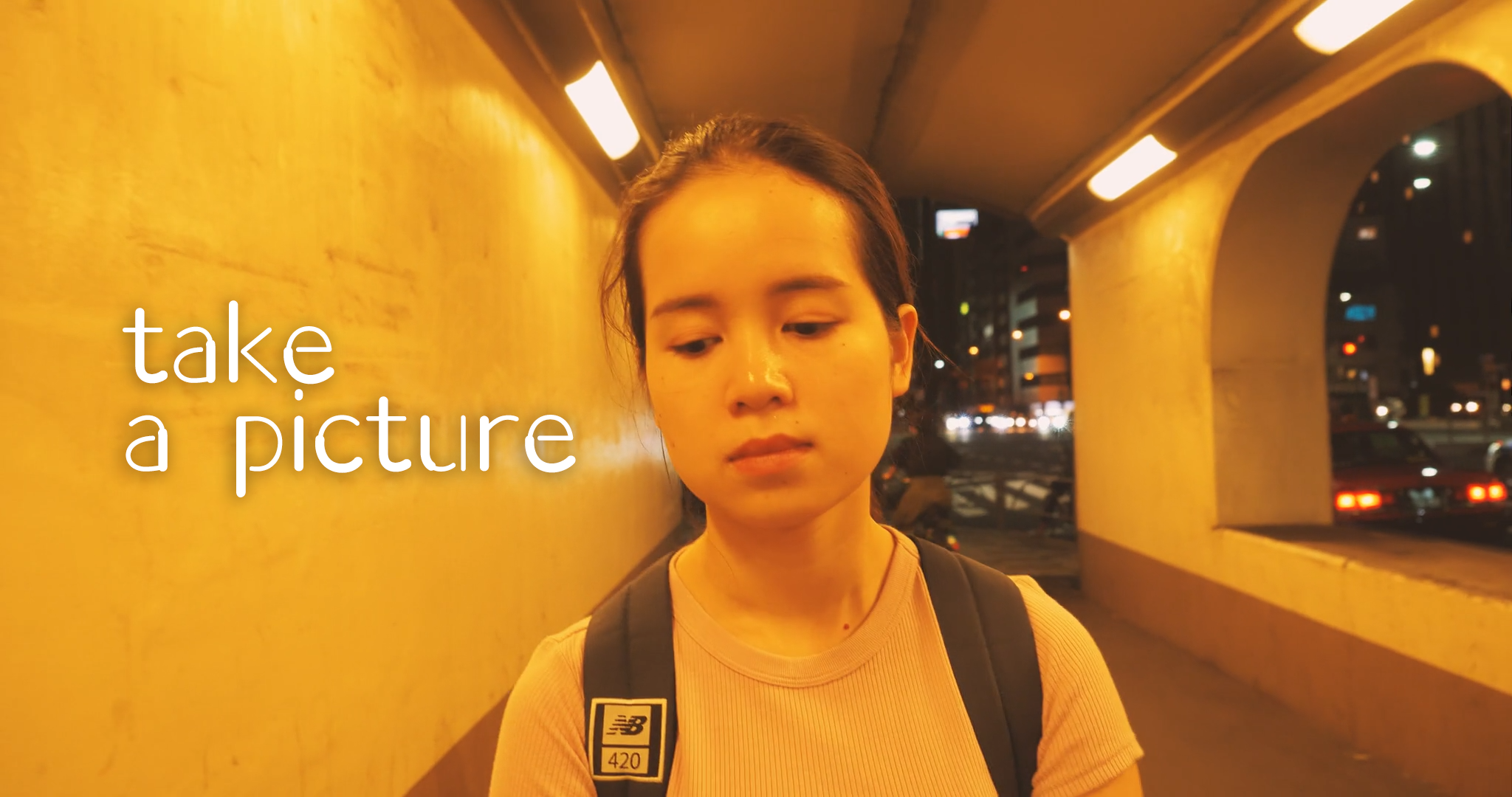 夜の都市の歩道橋で女性がカメラを見ずに立っている。背景には車のヘッドライトと街の照明が見える。