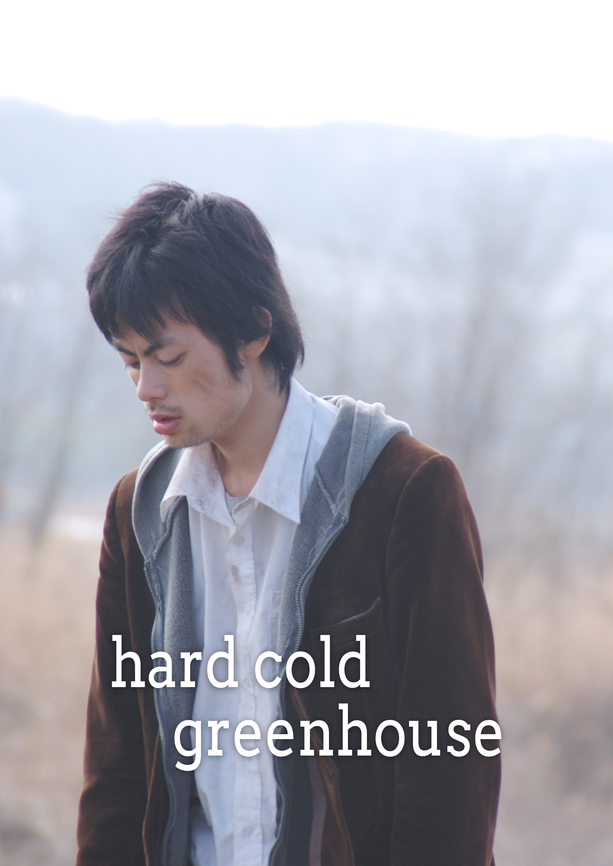 白いシャツと茶色いジャケットを着た男性が外で立っている。背景はぼやけている。