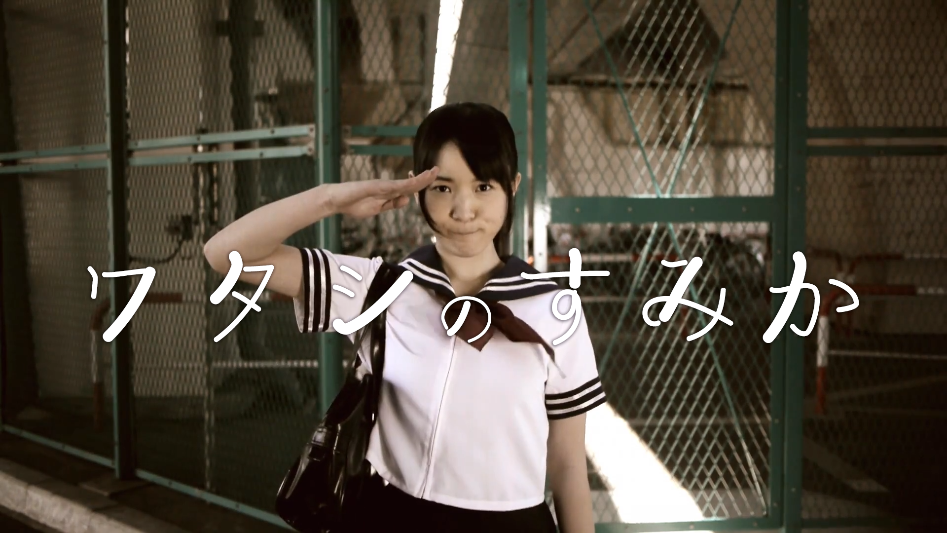 制服を着た少女が敬礼している。背景はフェンスや運動場のような場所。