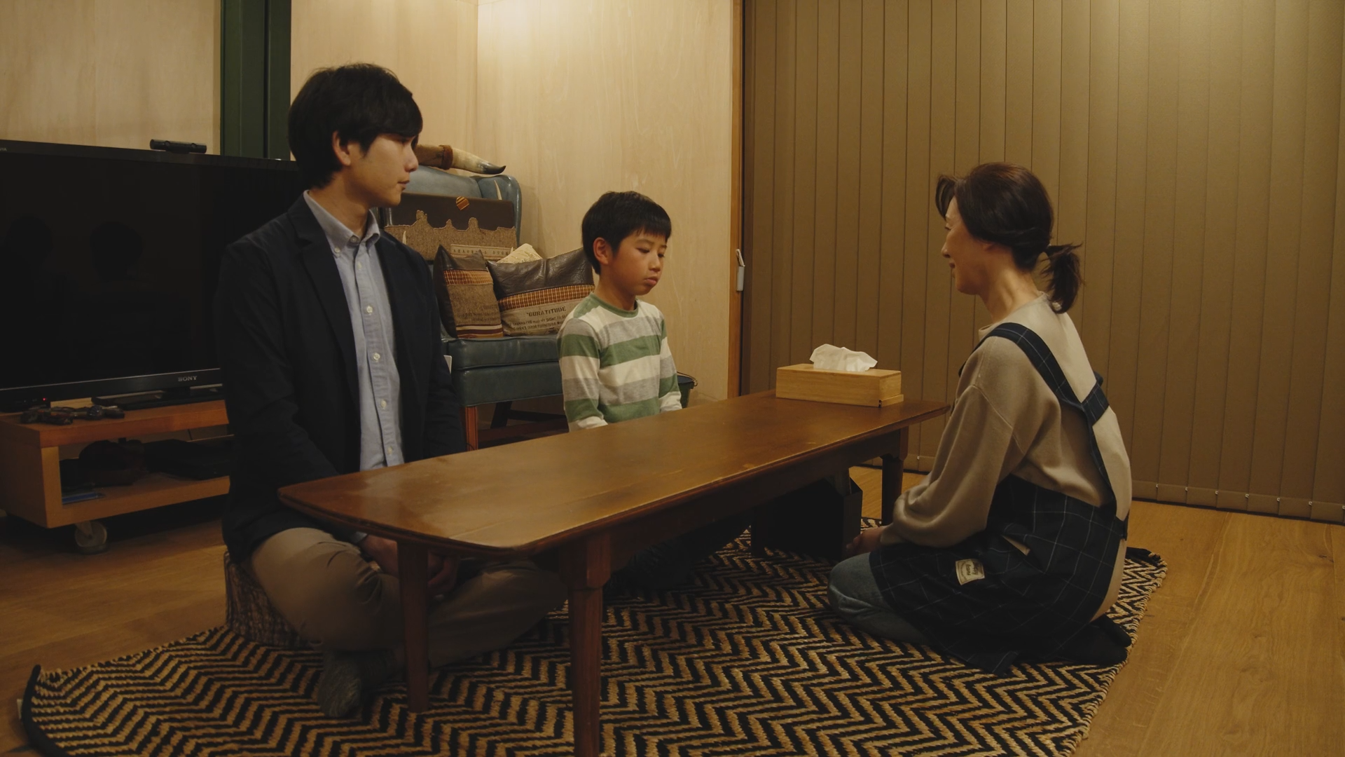 三人の家族が座って会話しているリビングルームのシーン。