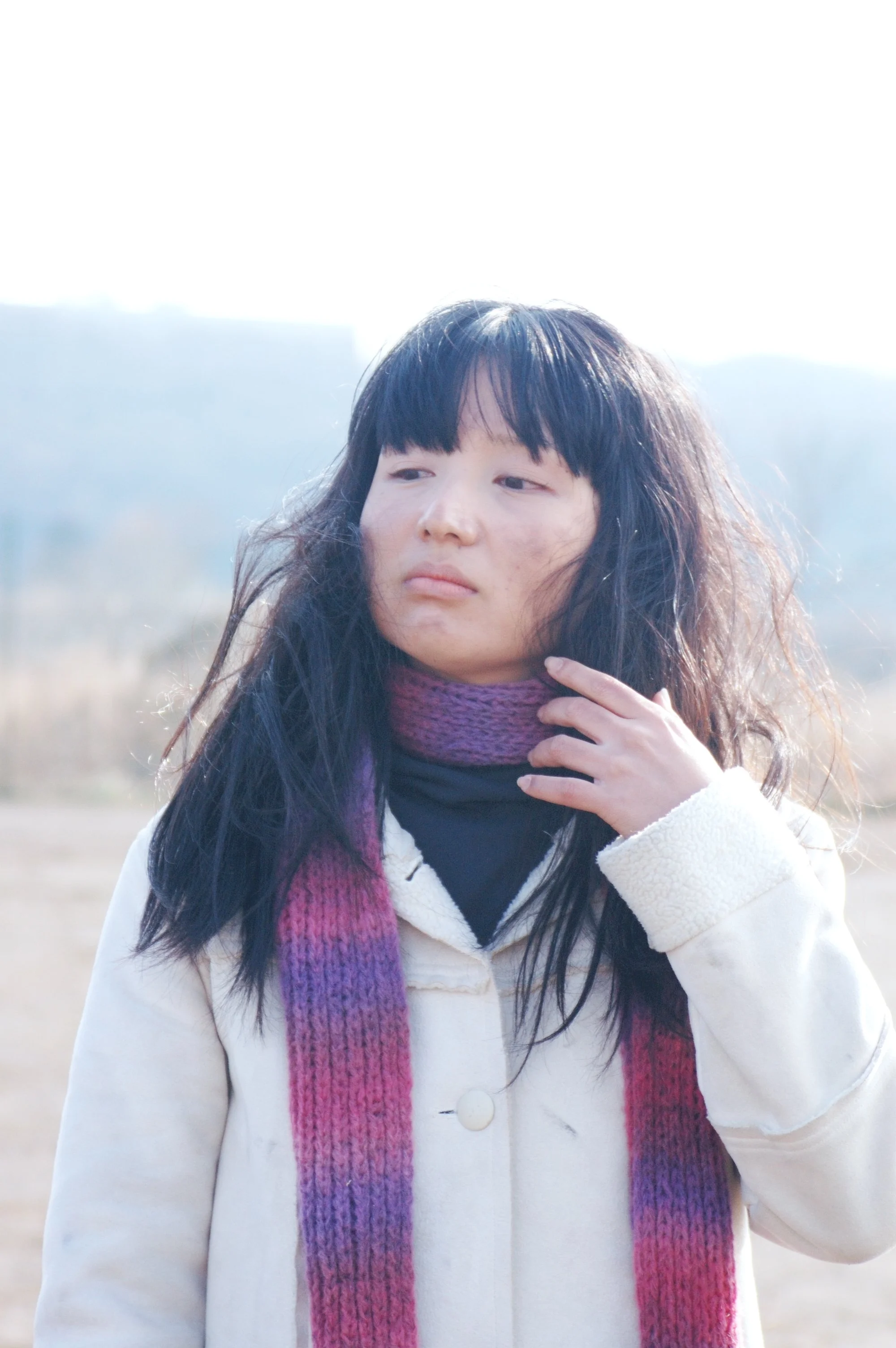 冬の野原で立っている若い女性、黒髪で苛立たしい表情で顔を見せ、紫とピンクのニットマフラーを着用しています。