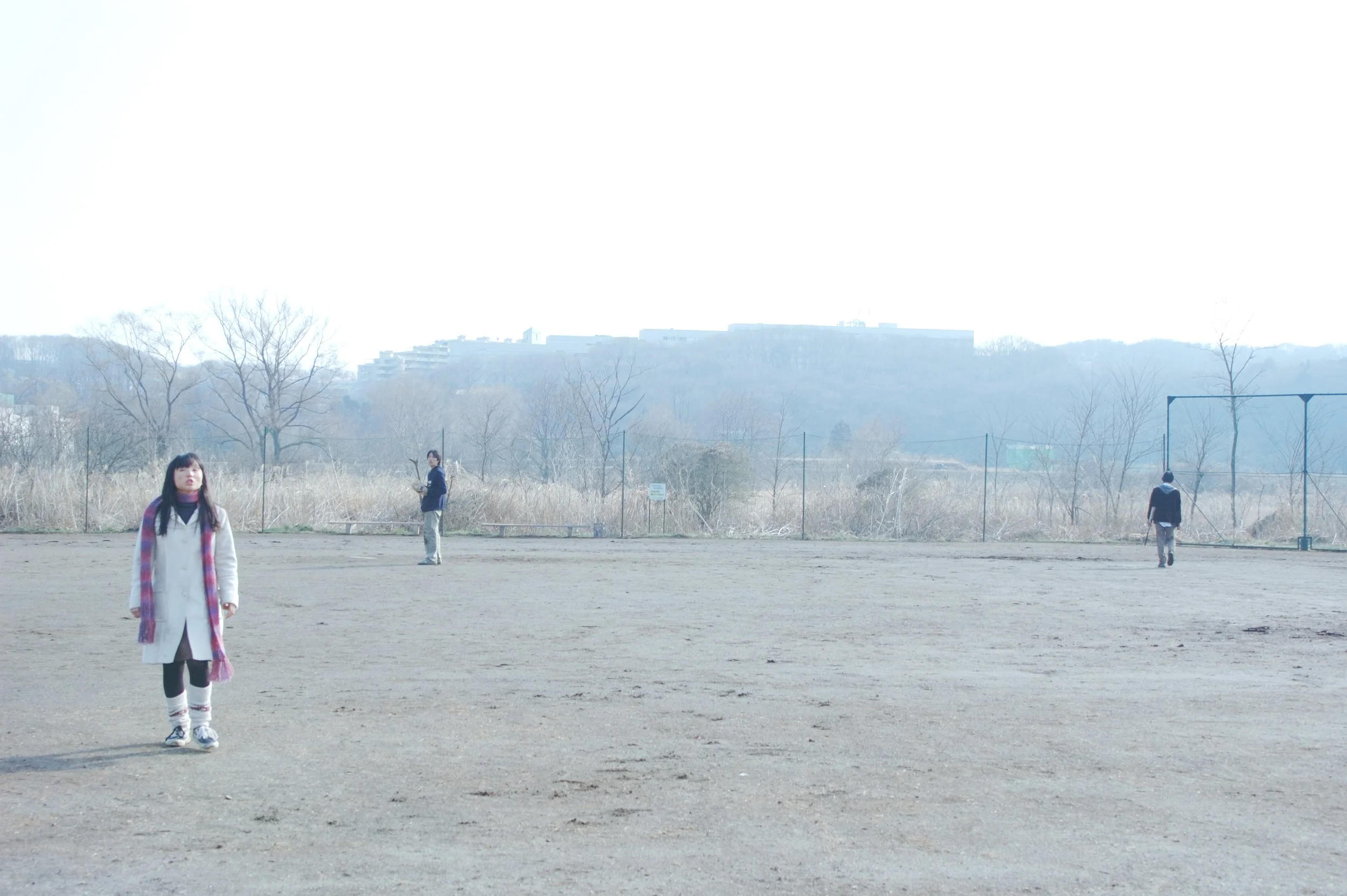 広い運動場で三人の人が離れた場所に立っている。背景には木と遠くの建物や山が見える。