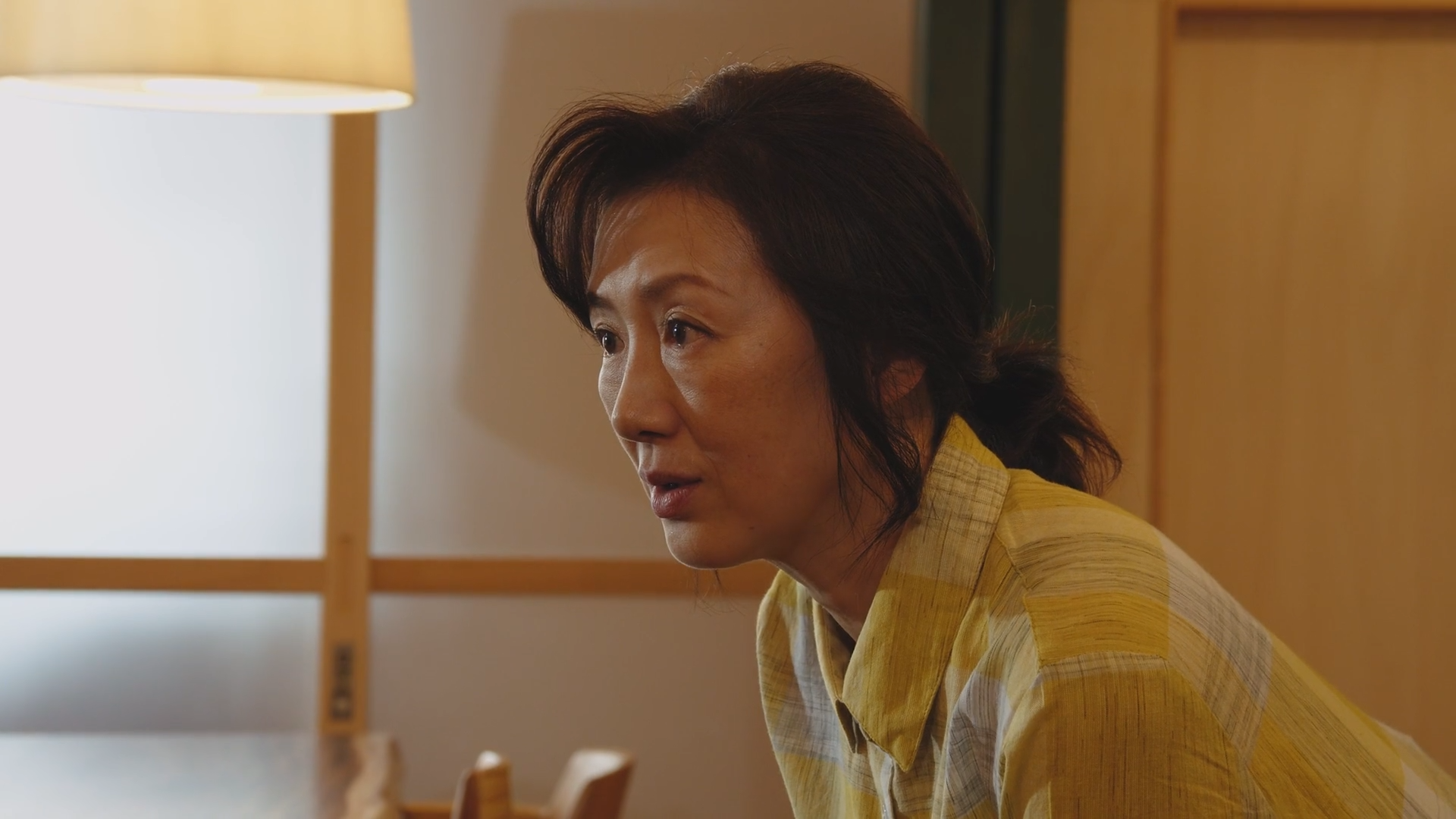 黄色いストライプのシャツを着た中年の女性が座っている。背景には木製のドアや間接照明が見える。
