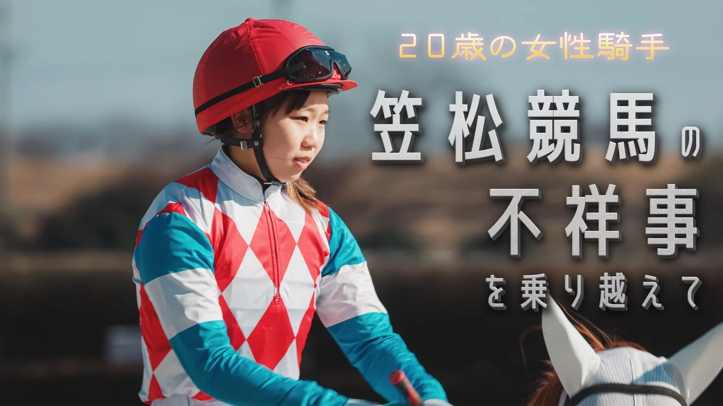 女性騎手がヘルメットとレースジャケットを着て馬に乗る様子。背景はぼやけた風景。