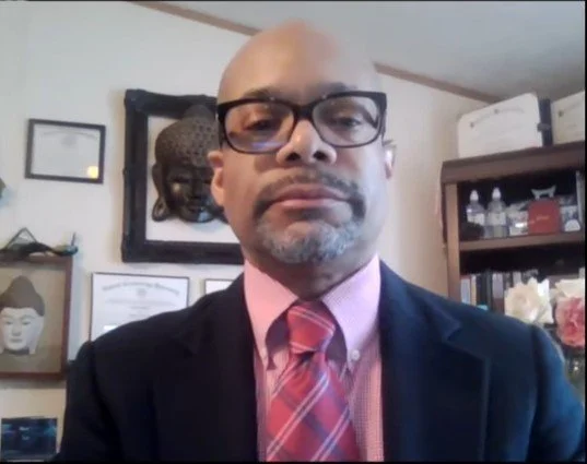Man in suit and glasses taking a selfie in a room with framed artwork and books on shelves.