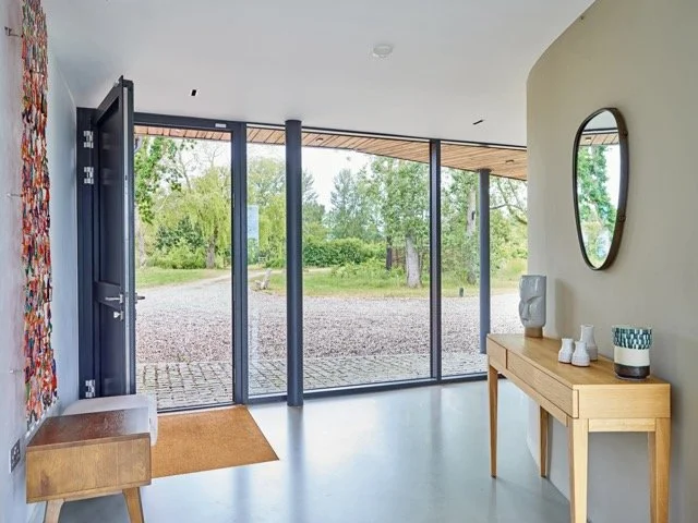 Modern entryway with a wooden console table, a round mirror, and decorative vases, leading outside through large glass doors to a gravel pathway and greenery.