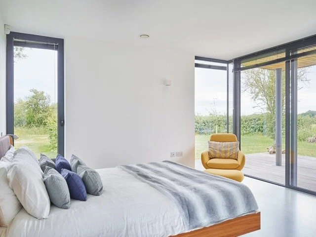 Modern bedroom with white walls, large windows showing greenery, a bed with white and gray bedding, and a yellow armchair near sliding glass doors.